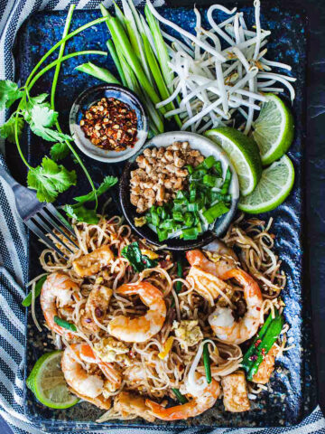 Shrimp Pad Thai on a blue plate with lime wedges, green onion, chilis and peanuts on the side.