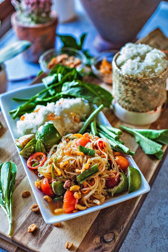 A plate of Thai papaya salad with chicken wings, veggies, pork rind and sticky rice around it.