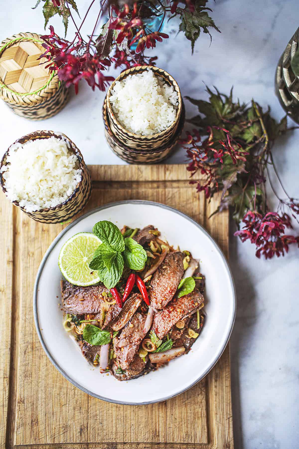 Nam Tok Beef waterfall salad on a plate on a cutting board with sticky rice baskets.