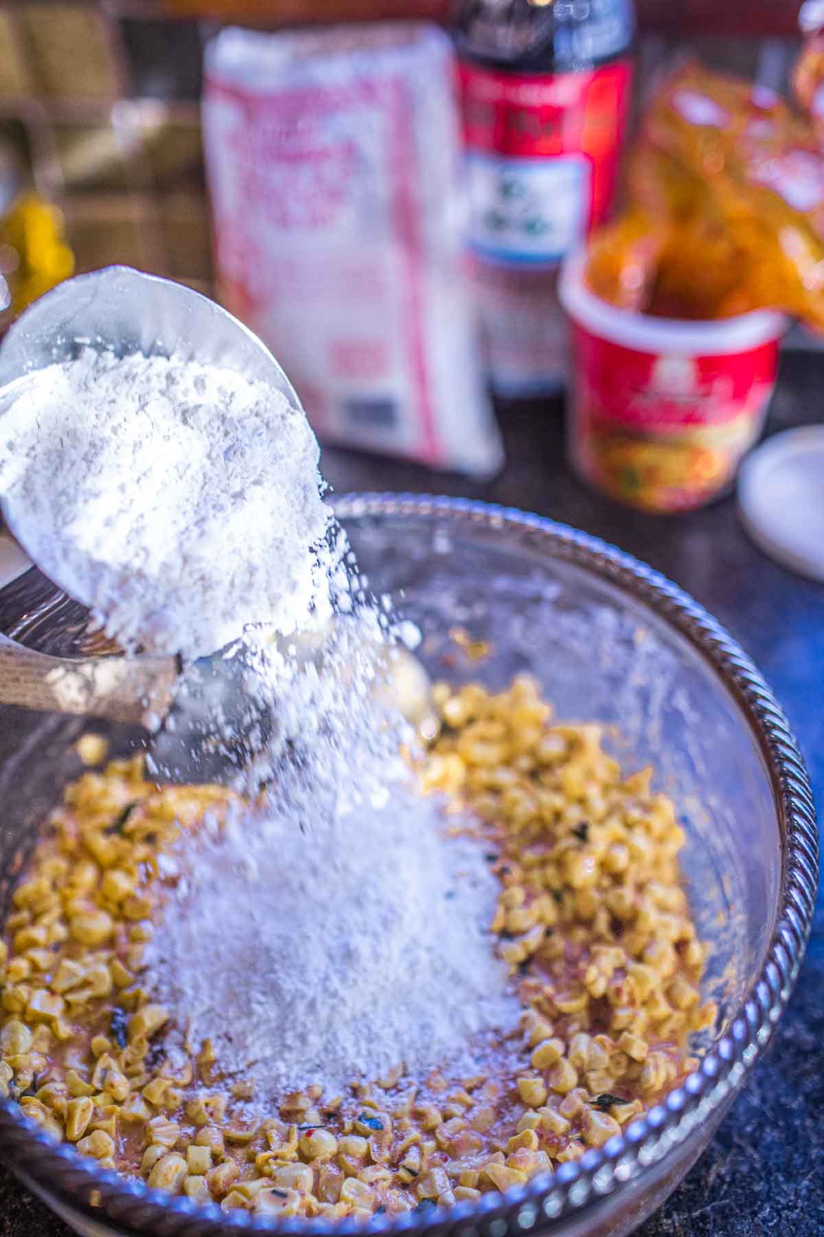 pouring flour into corn fritter batter in a glass bowl with spatula