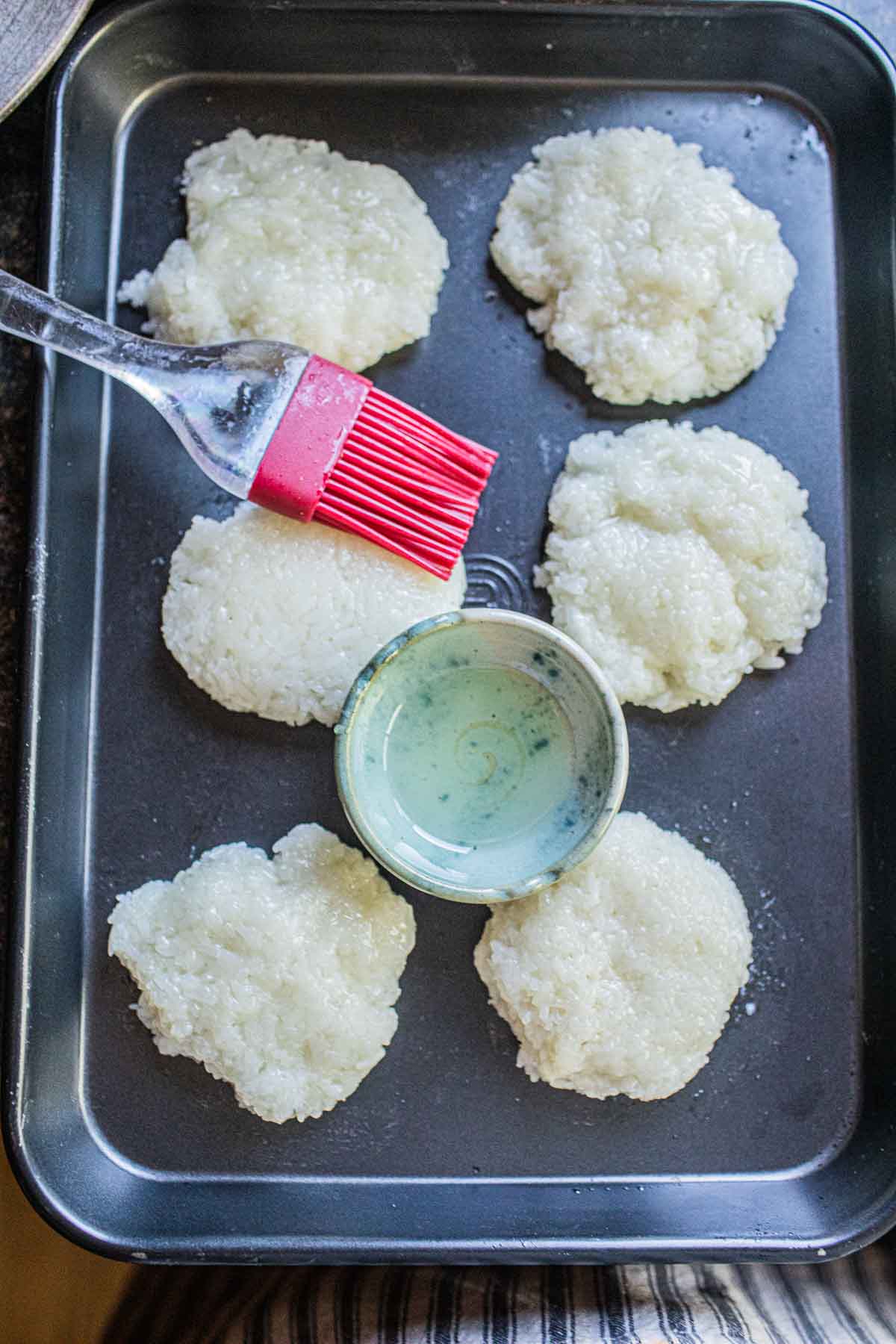 Khao Jee patties on a baking tray with oil and brush.