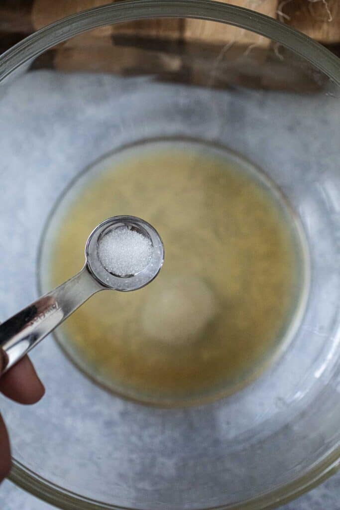 Sugar in a measuring spoon over a bowl of dressing.