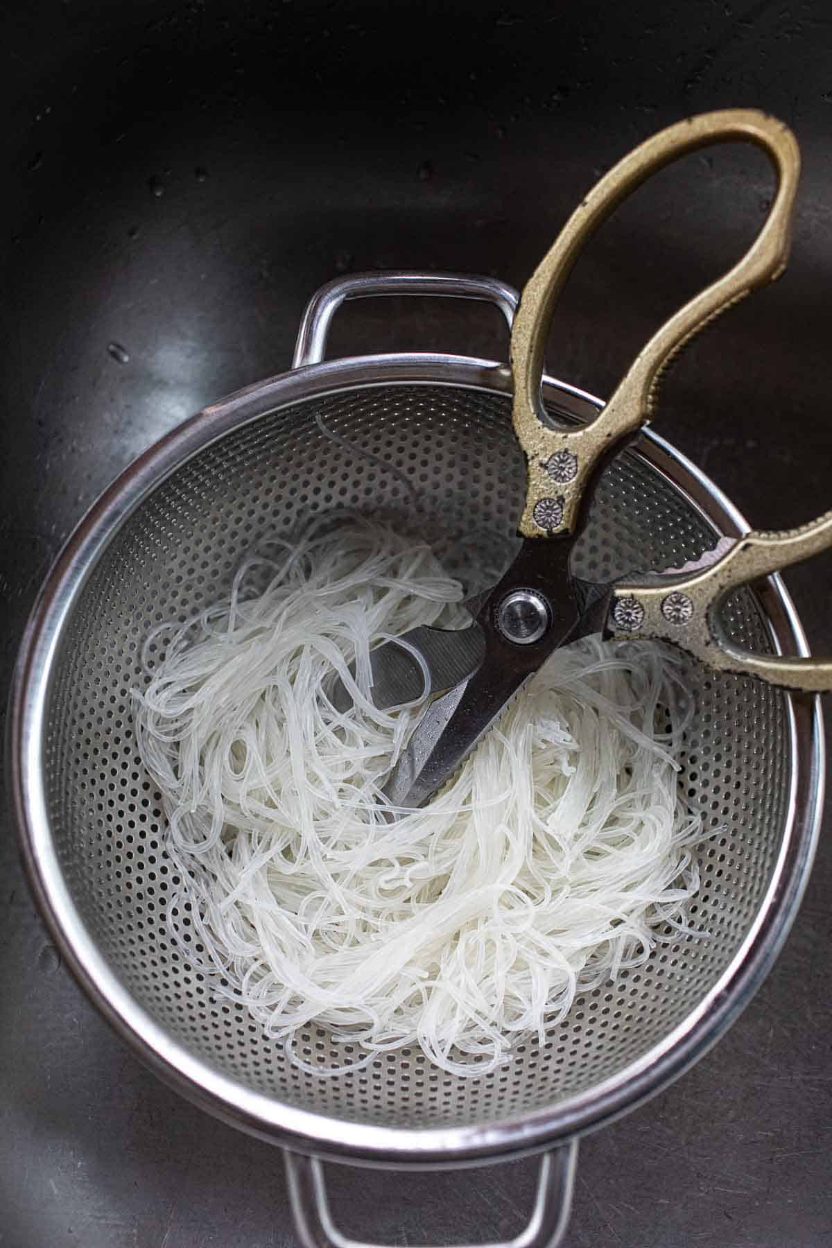 Scissor cutting glass noodles in a colander.