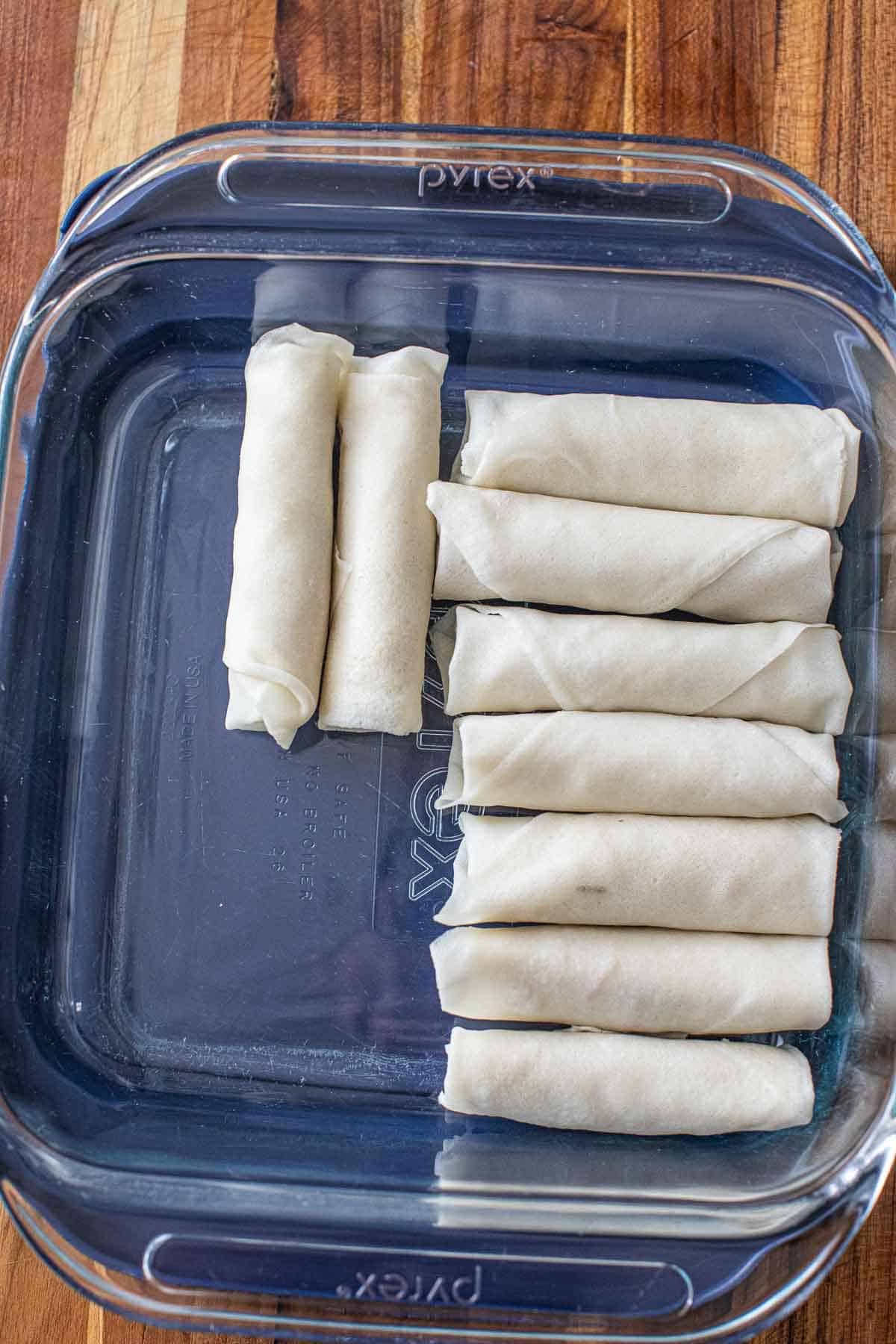 Uncooked wrapped egg rolls in a glass container. 