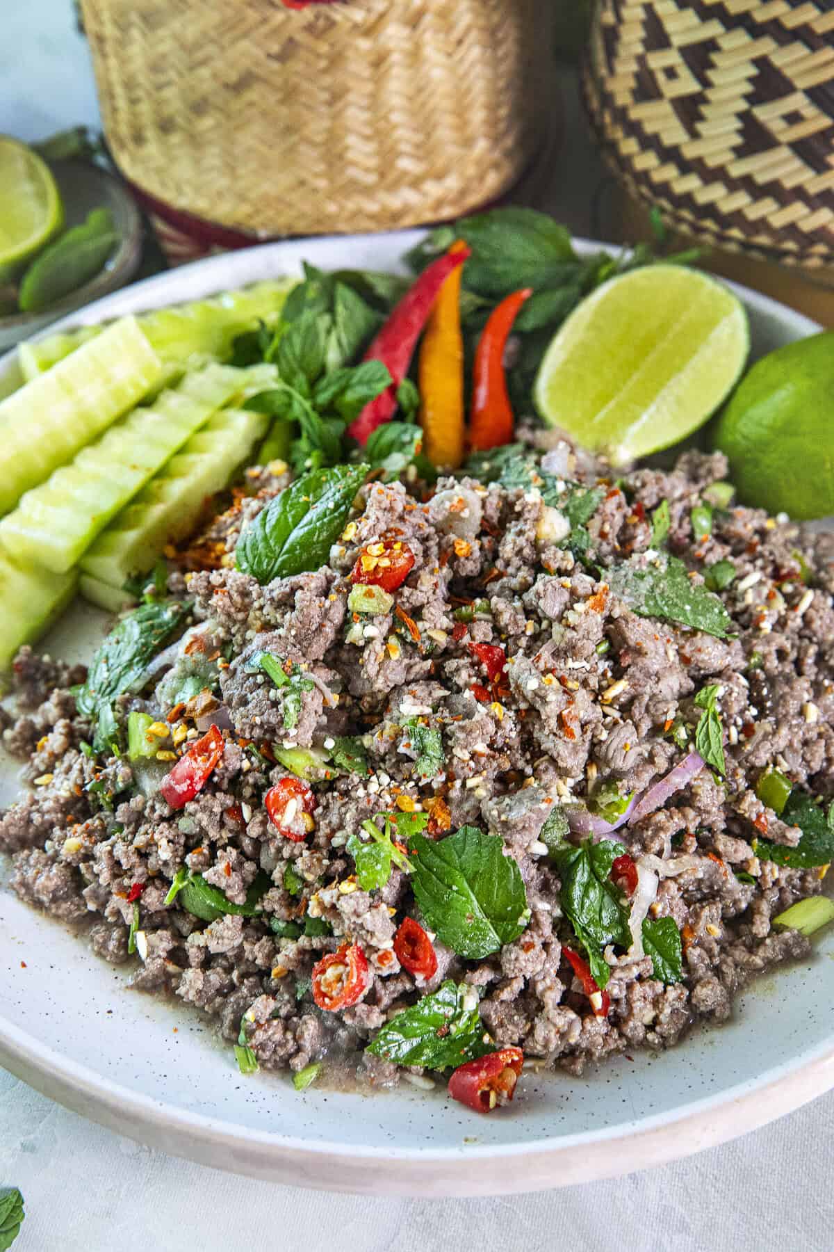 Ground meat Beef laab on a platter with garnishes of lime wedges, cucumbers and sticky rice baskets.