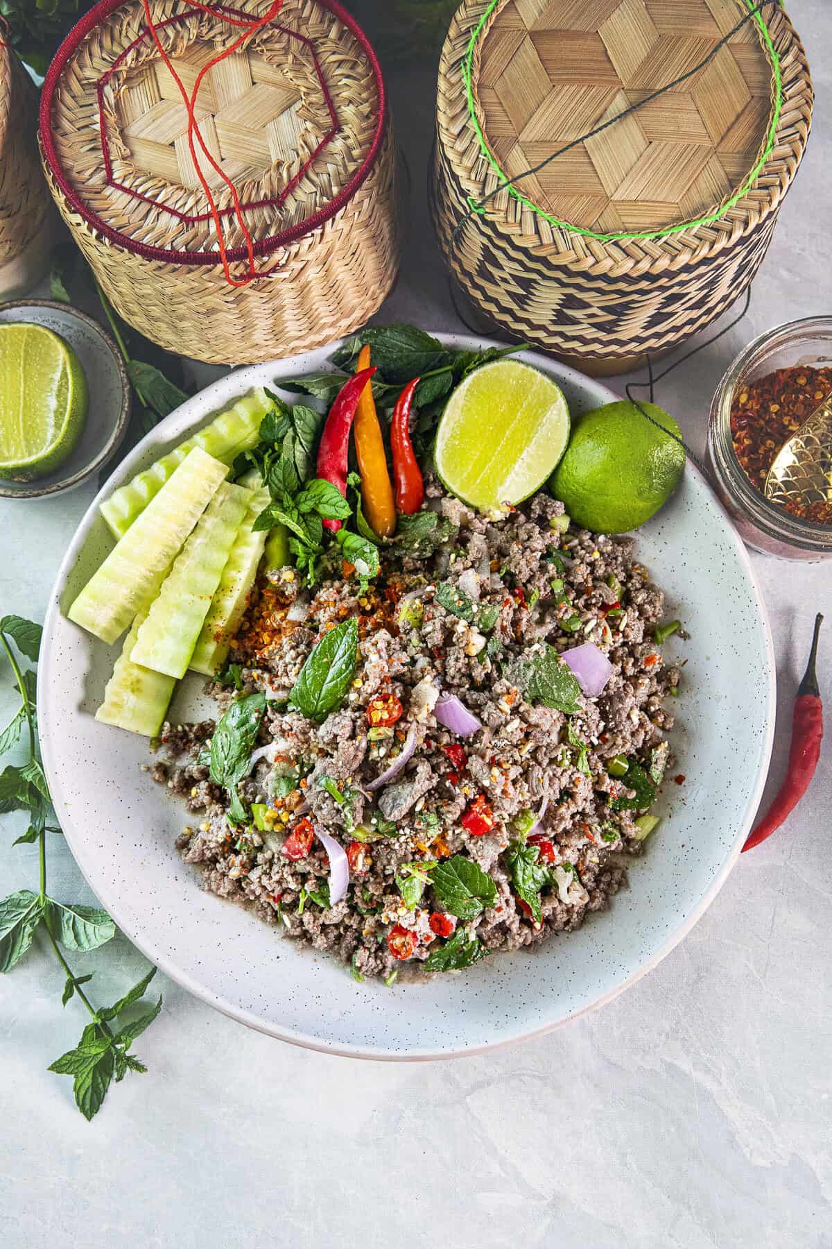 Authentic Thai Beef Laab in a platter with fresh herbs and cucumber garnishes and sticky rice baskets on the side.