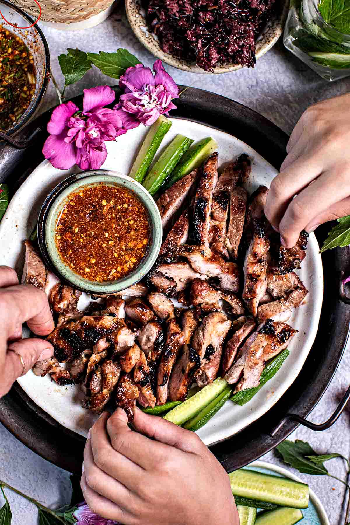 Fingers grabbing Thai grilled pork neck from a platter.