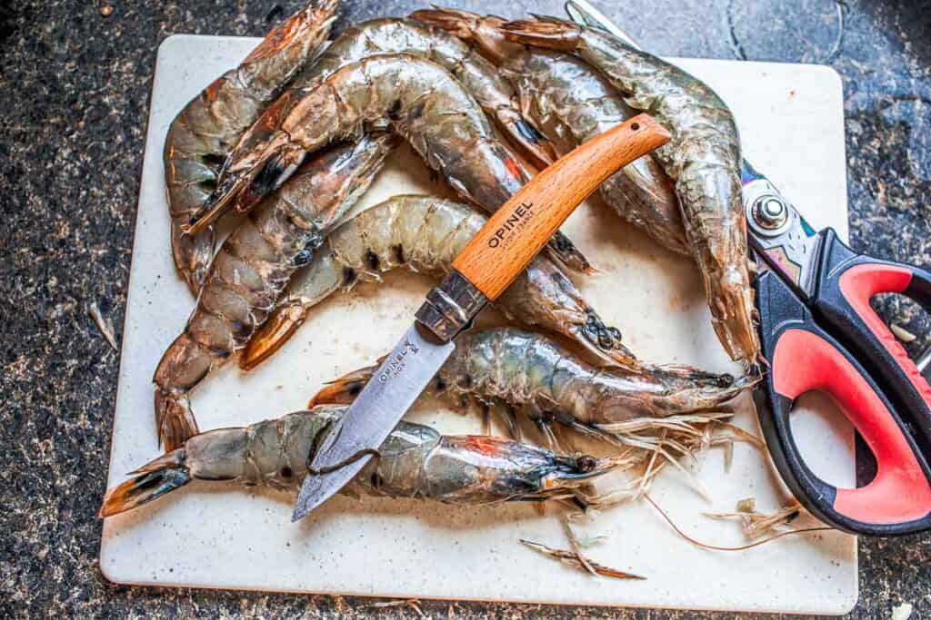 shrimp with shells on a cutting board with a knife deveining. 