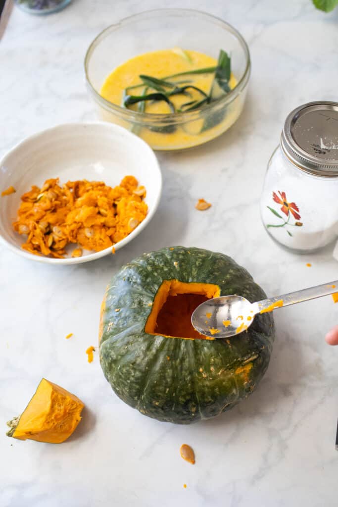 A spoon over Carved hollow up green pumpkin next to a bowl of fibers and seeds.