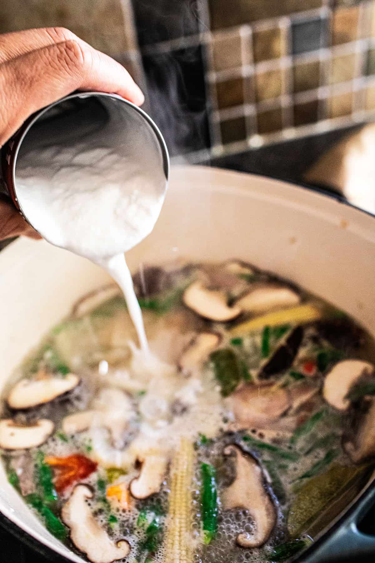 Canned coconut milk pouring into tom kha soup pot. 