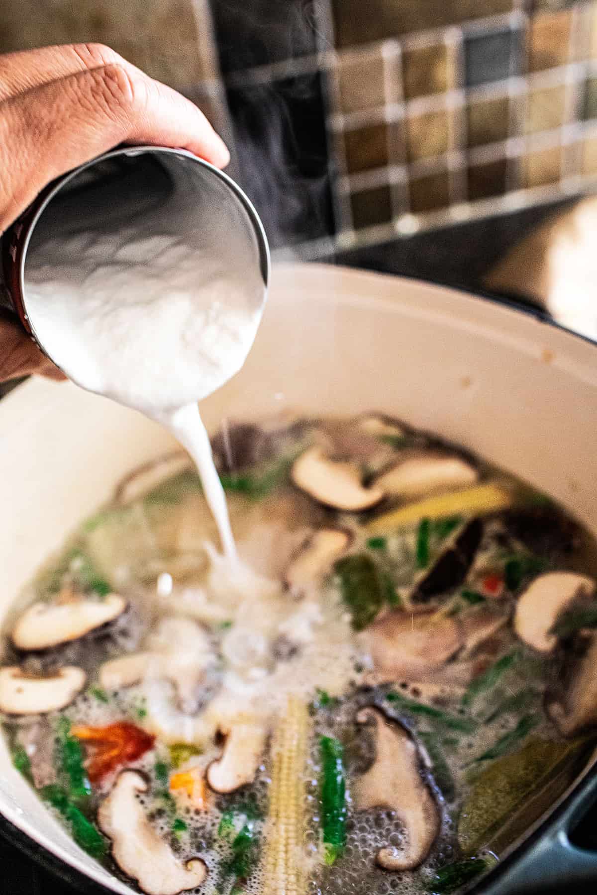 Canned coconut milk pouring into tom kha soup pot. 