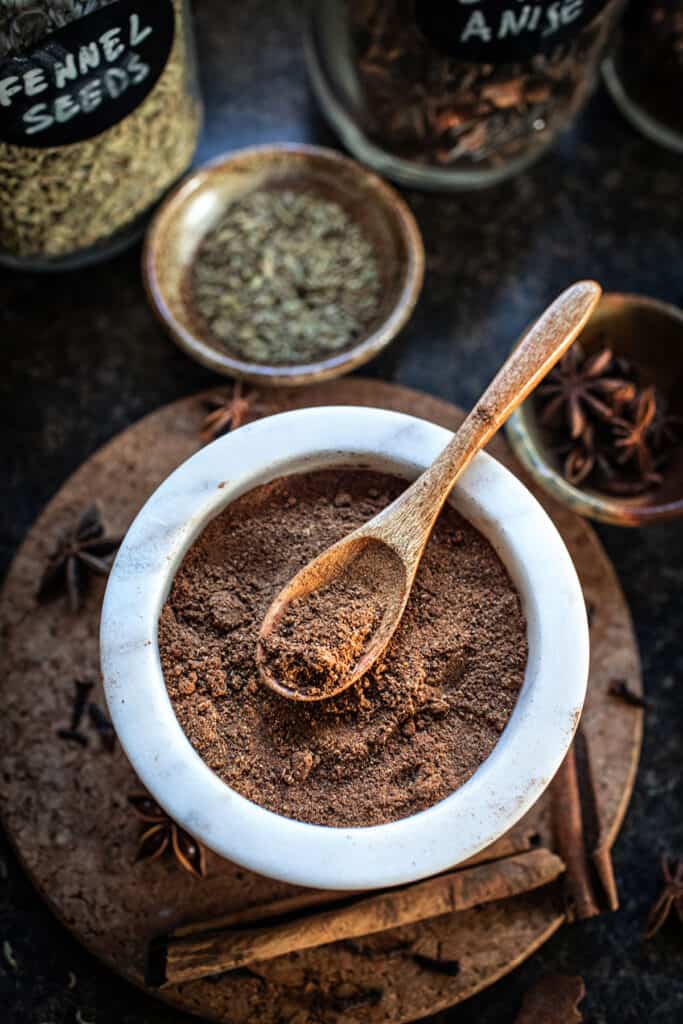 Spoon holding 5 spice powder in a white bowl.