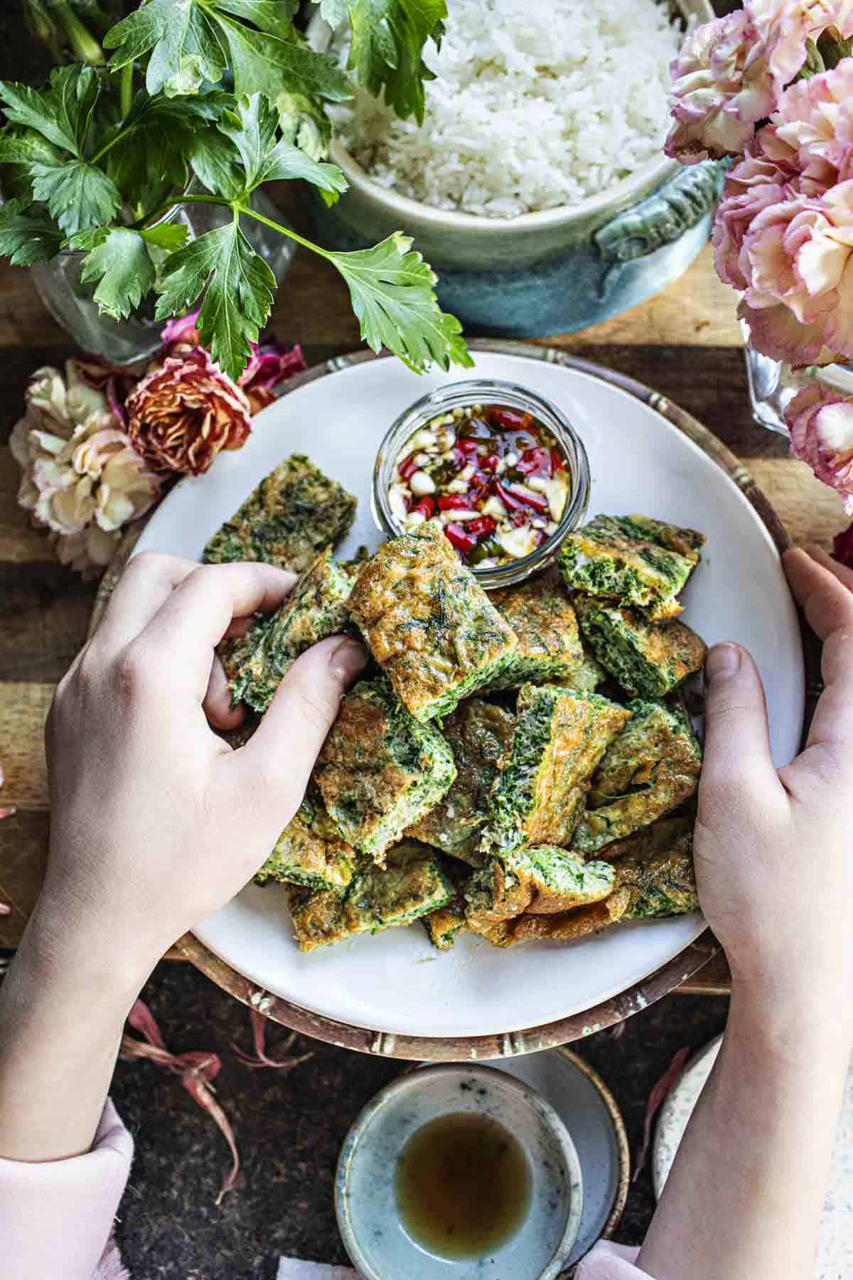 Two small hands holding Thai Cha Om Omelette squares on a plate. 