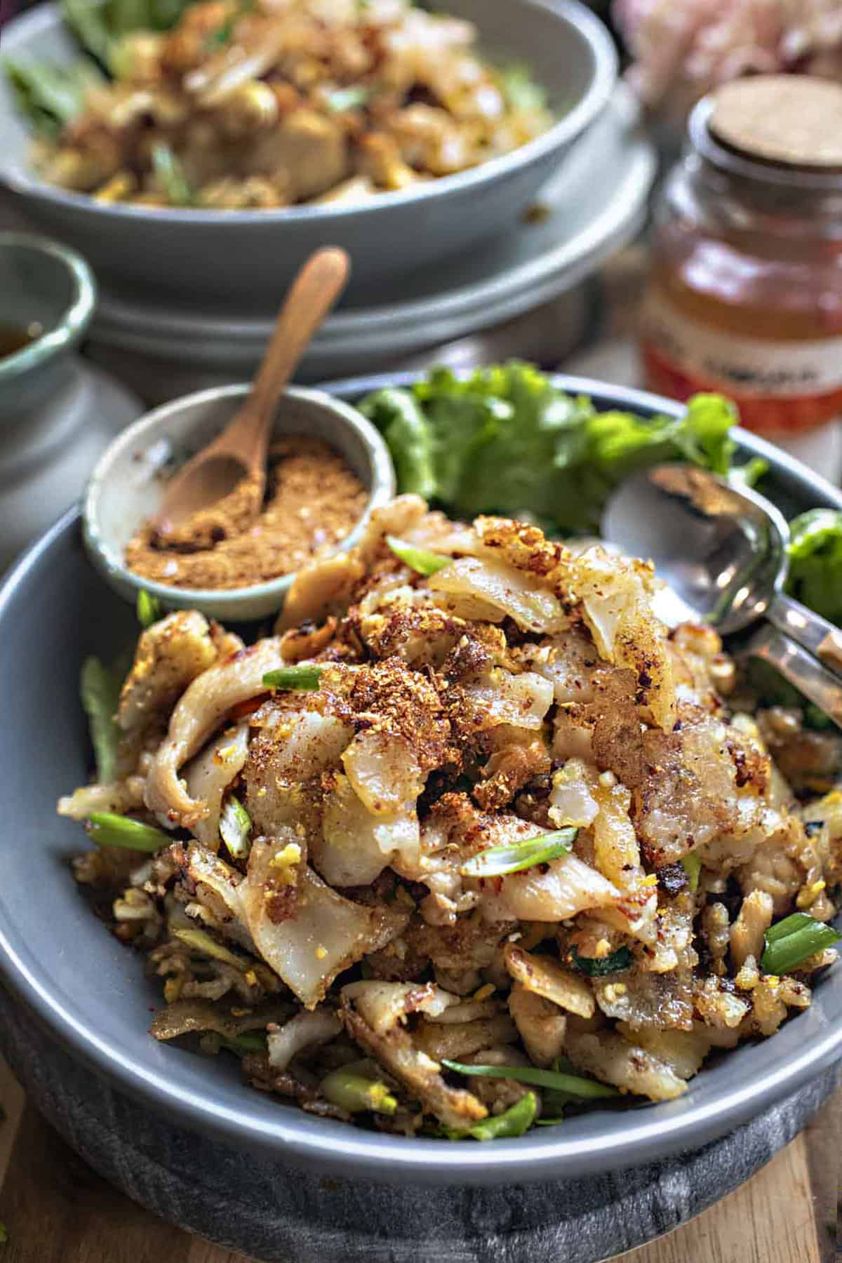 Wide rice noodles on a platter with chili flakes on the side. 