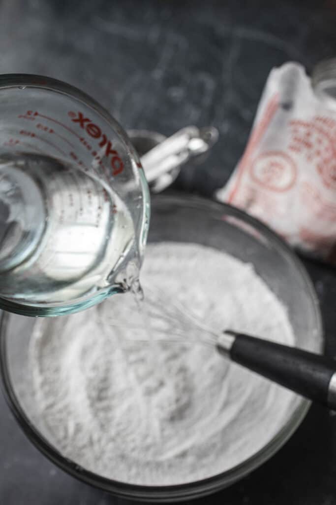 water pouring into Flour and whisk in a glass bowl on the table. 