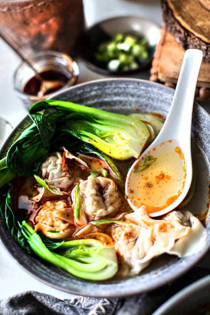 A spoon inside a Pork wonton soup in a bowl with bok choy.