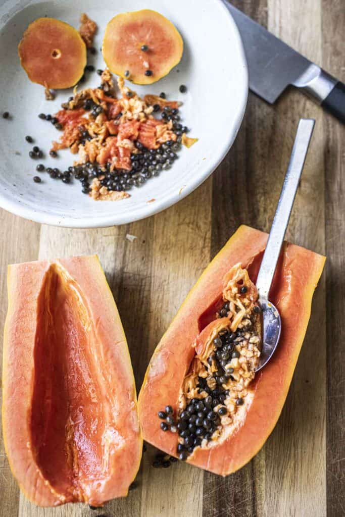 a spoon scrapping ripe papaya in 2 halves on a cutting board. 