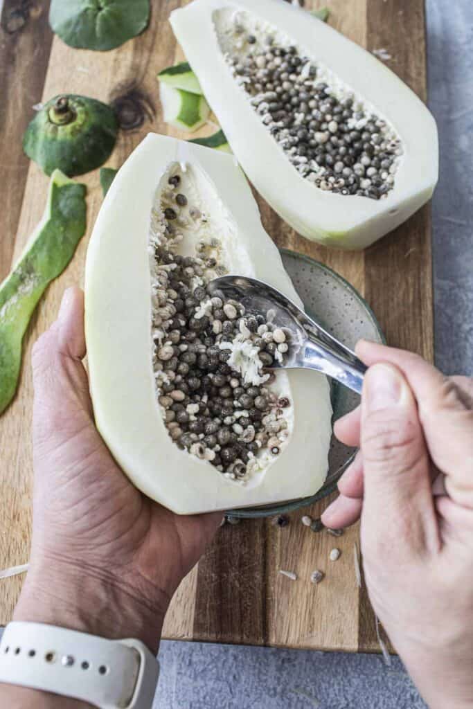 A hand holding half of green papaya with a spoon scraping seeds. 