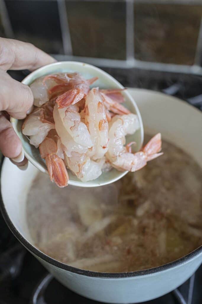 Shrimp in a bowl pouring into a soup pot on the stove. 