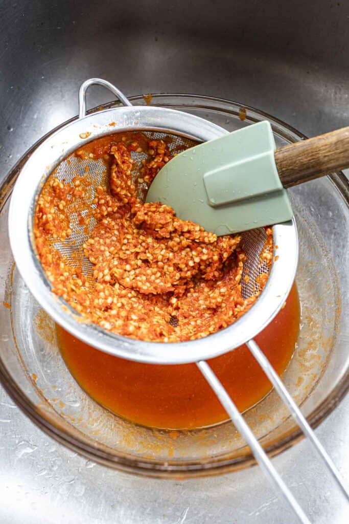 Spatula pressing into Thai hot sauce in a strainer into a glass bowl. 