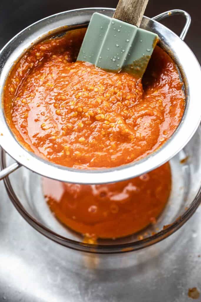 Spatula pressing into Thai sriracha sauce in a strainer into a glass bowl. 