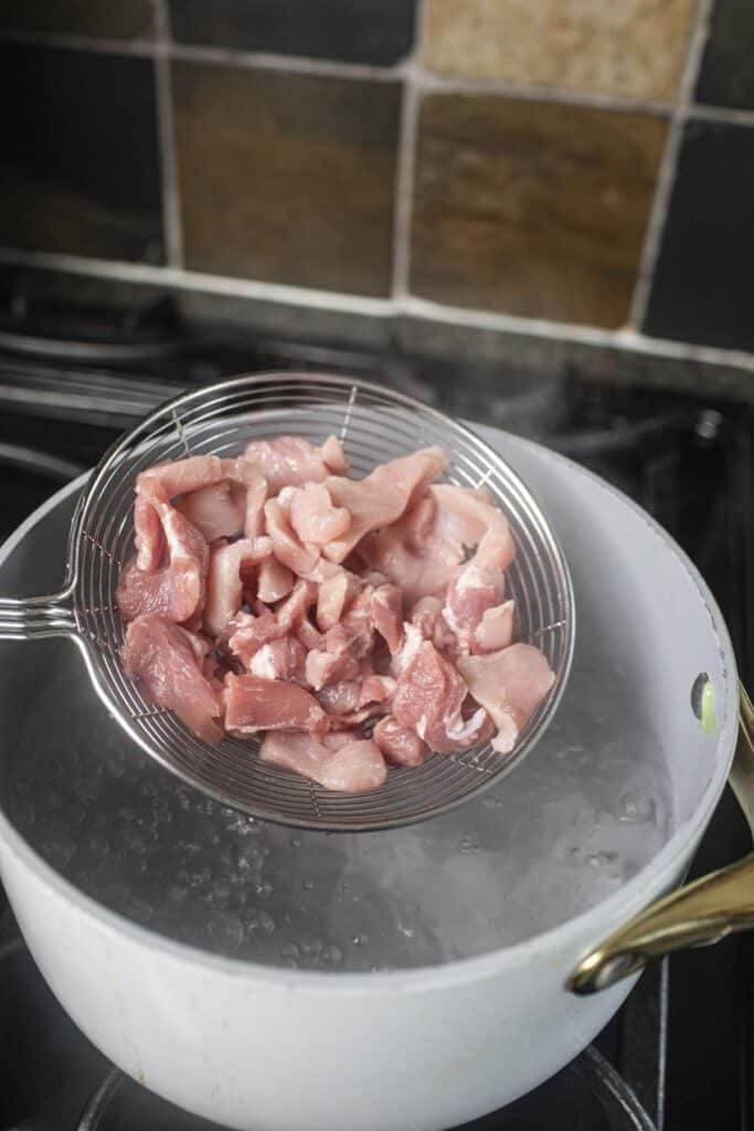 Wire mesh skimmer with raw pork slices over a pot.