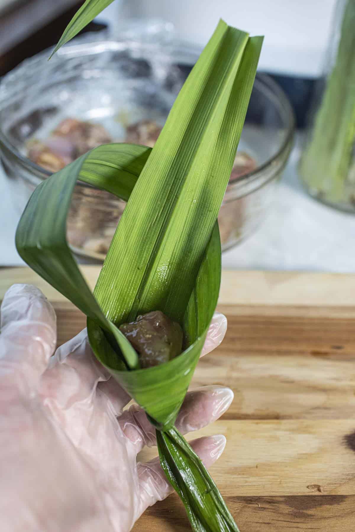 Gloved hand holding wrapped pandan chicken in a leaf.