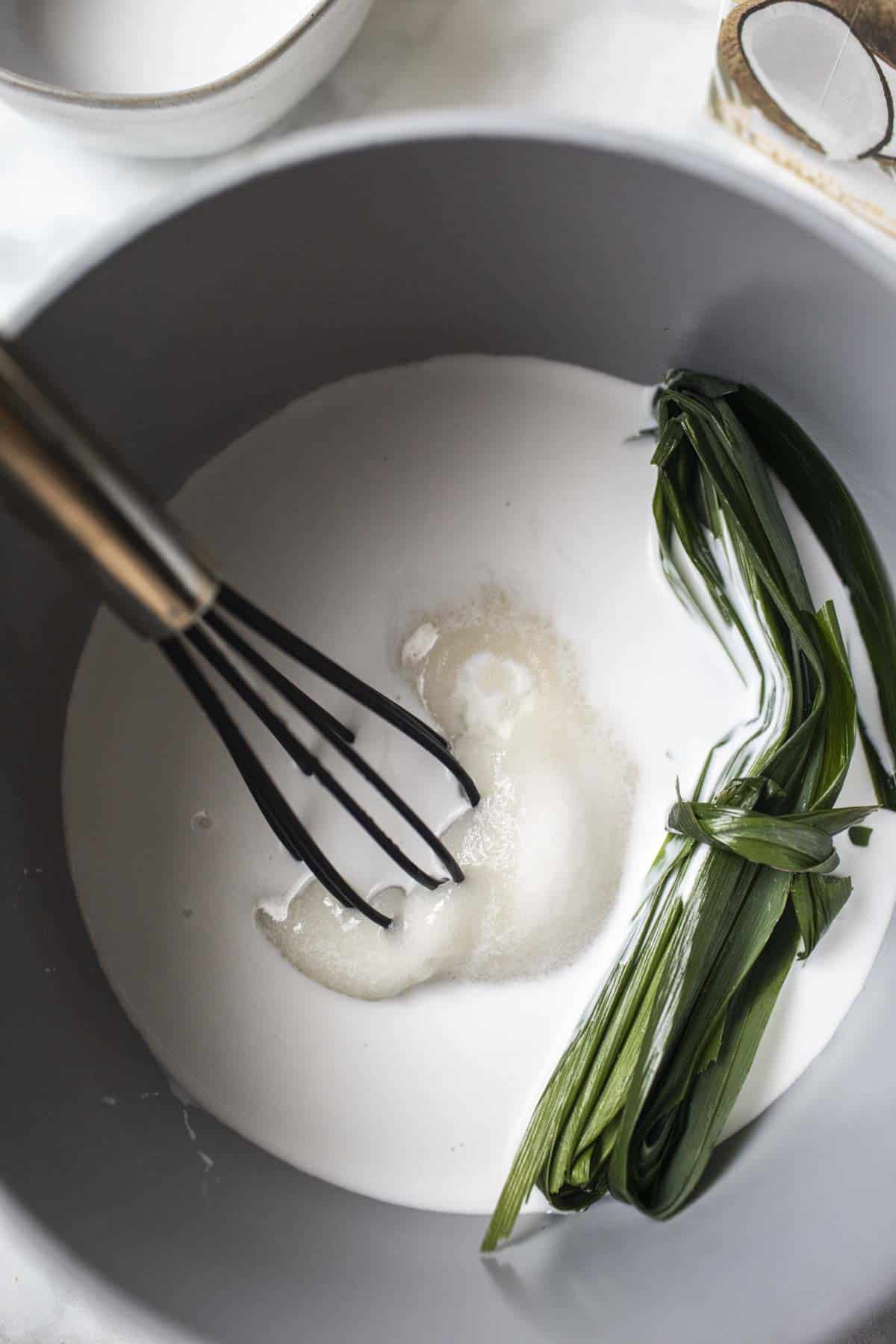 Sugar, coconut milk and pandan leaves in a pot with a whisk. 