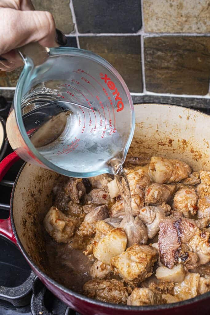 Water pouring over pork chunks with hung lay paste in a dutch oven.