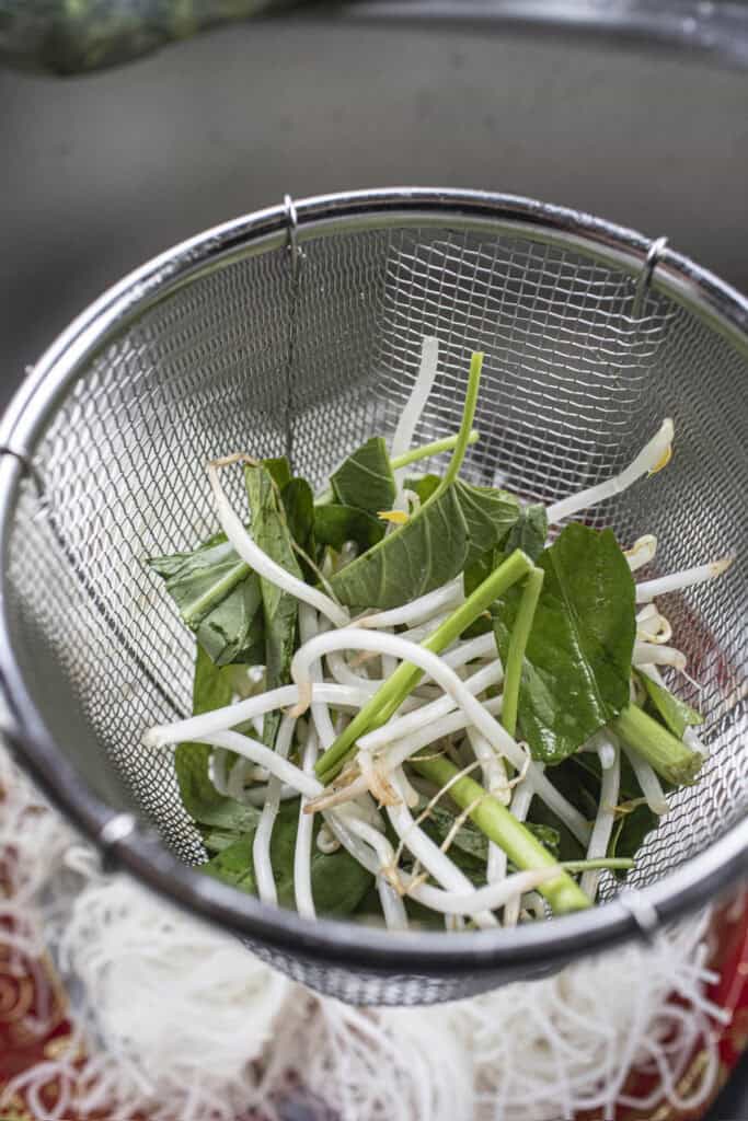 Bean sprouts and water spinach in a metal basket over a broth in a pot.  