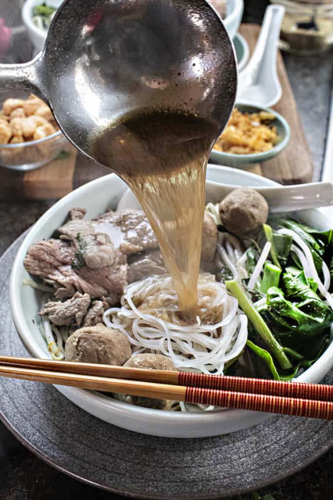 A metal laddle pouring broth into a bowl of noodles and meat and veggies. 