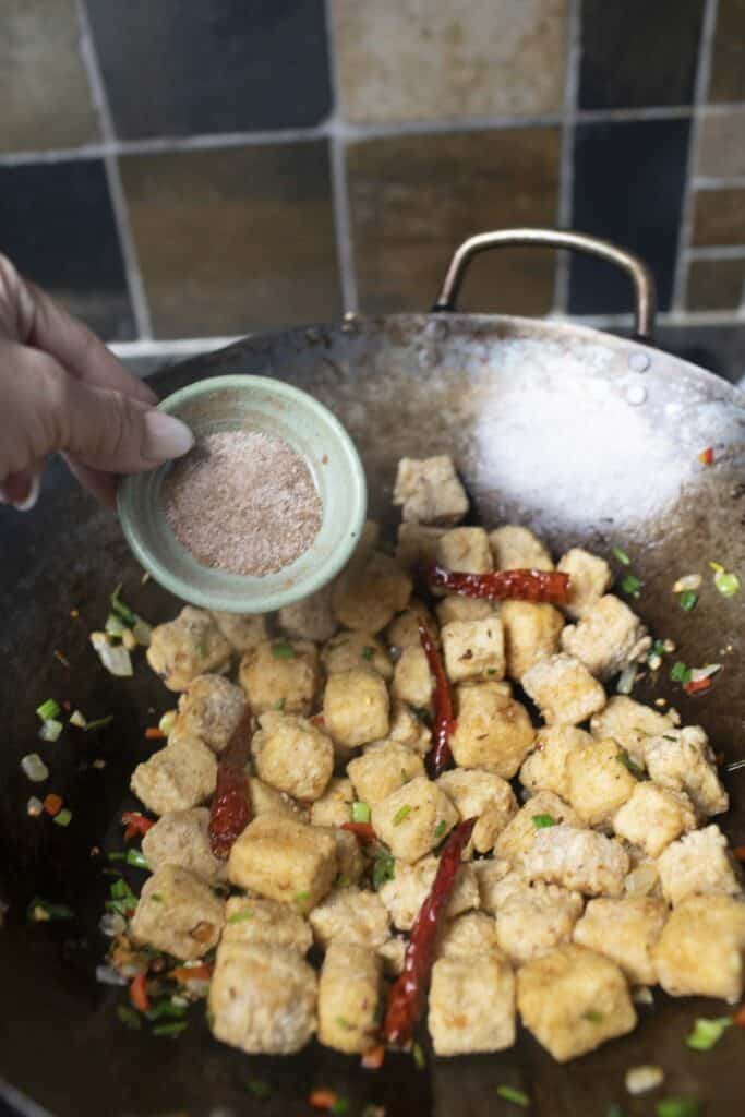 Five spice blends in a bowl, pouring into a wok full of salt n pepper tofu. 