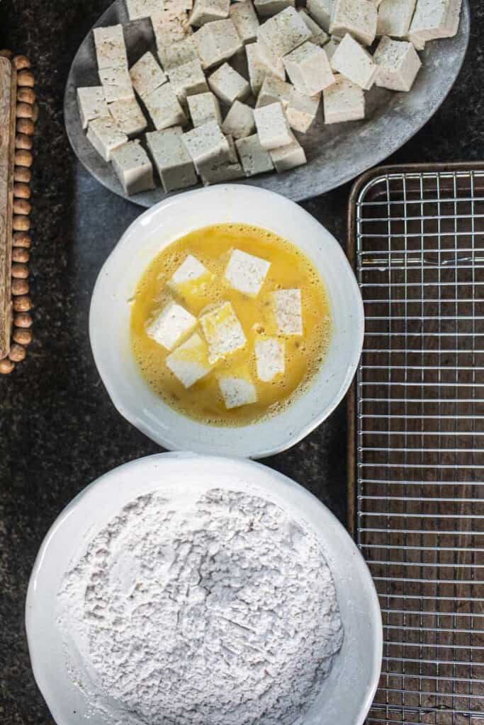  Tofu cubes in whisked egg next to a bowl of flour. 