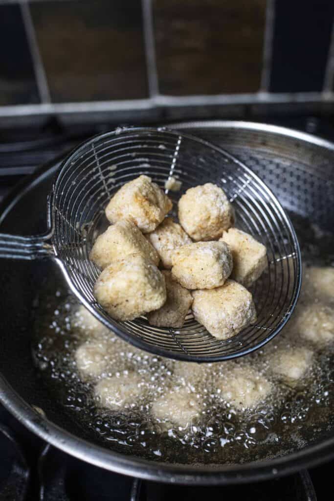 A wire mesh strainer lifting fried tofu from hot oil in a wok. 