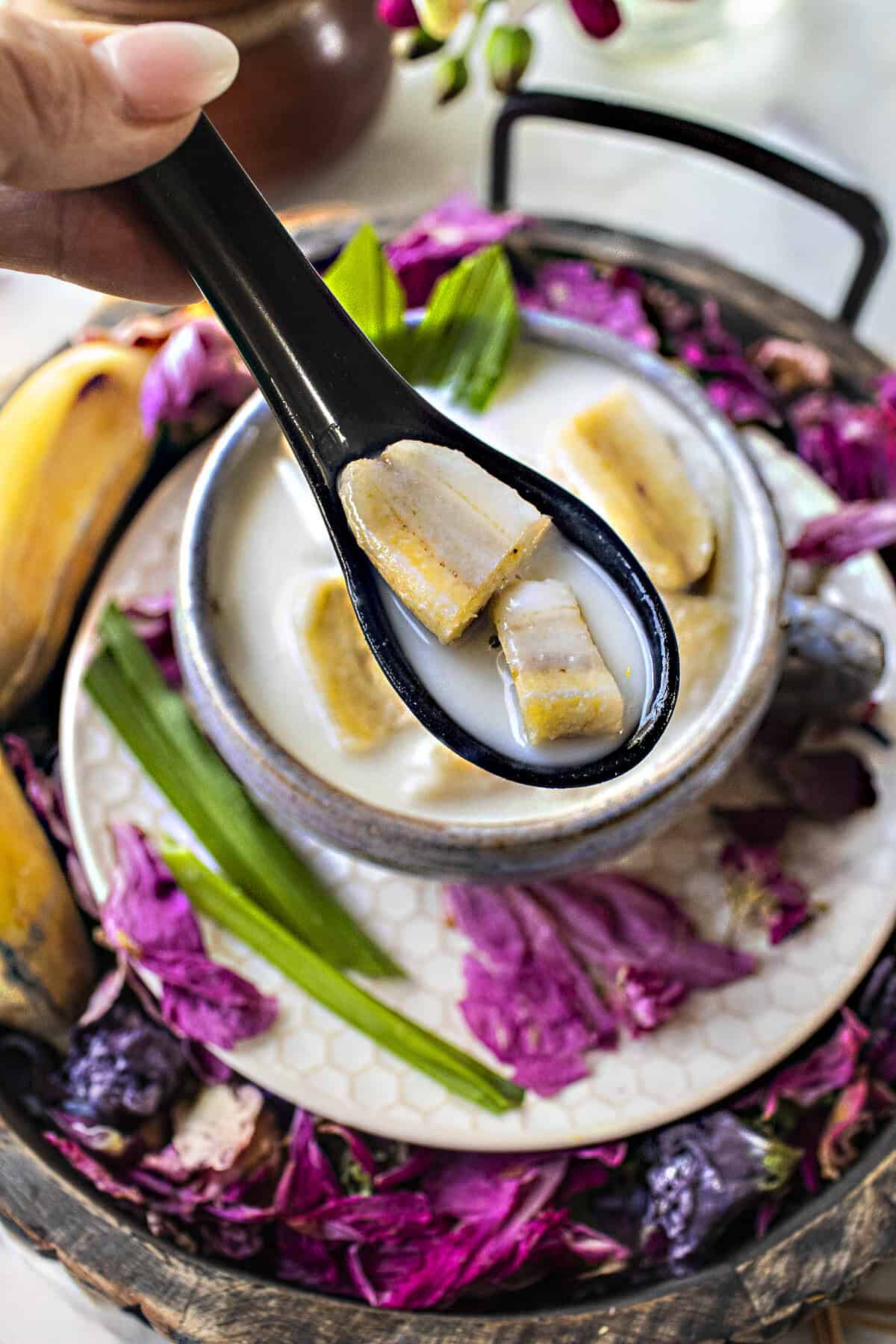 A spoon lifting kluai buat chi banana dessert in a bowl surrounded by pink flower pedals.