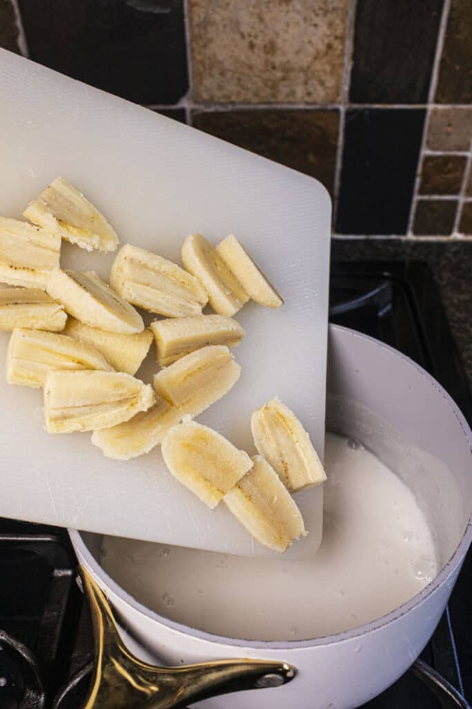 Sliced Thai banana on a cutting board, pouring into coconut milk in a pot. 