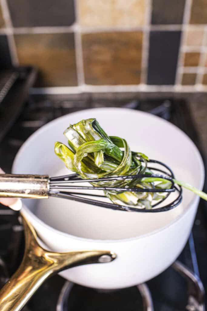 A whisk lifting pandan leaves in coconut milk in a saucepan. 