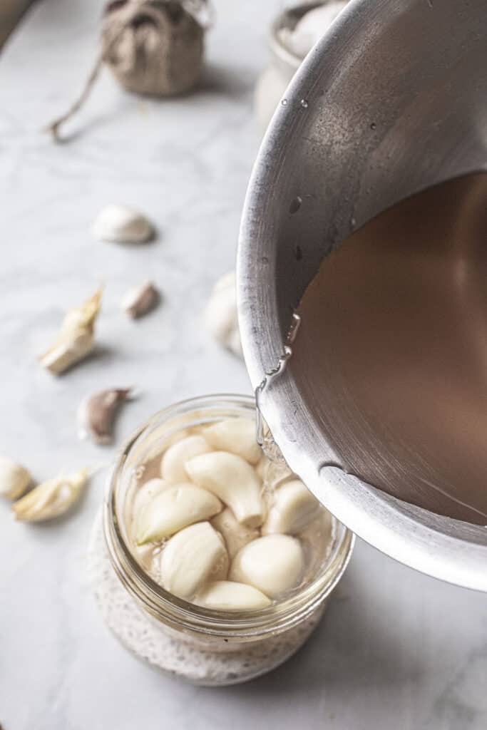 Brine pouring into a glass jar with garlic cloves. 