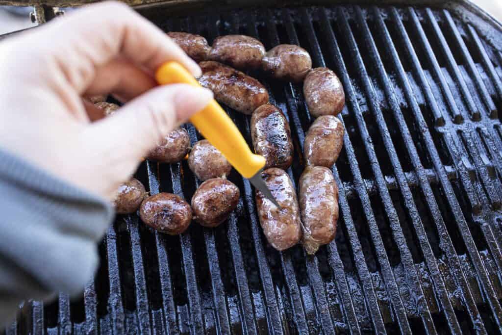 hand poking grilled sausages on a grill with a knife. 