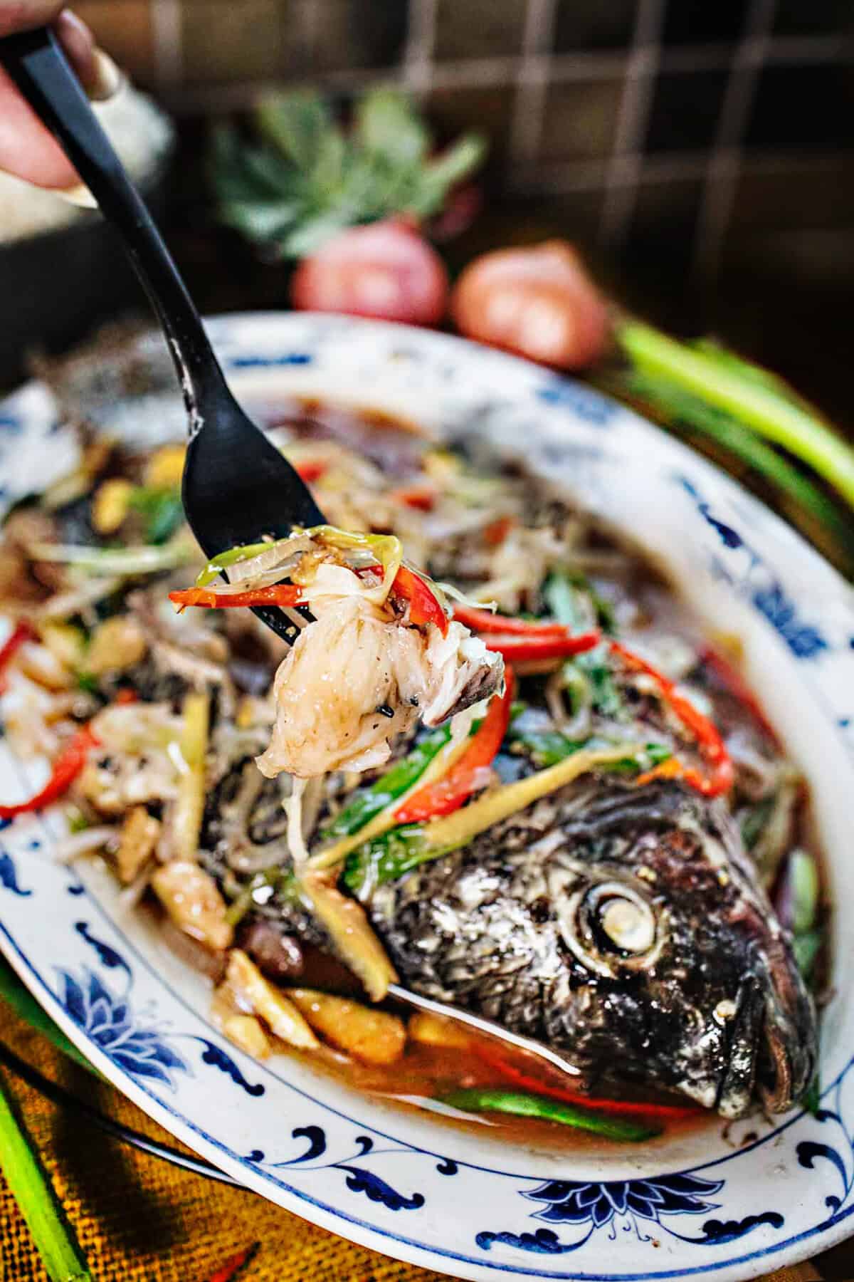 A fork lifting fish meat from a whole steamed fish with herbs on top on a platter. 