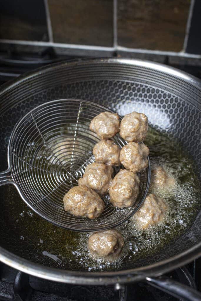 Wiremesh skimmer lifting meatballs from oil in a pot. 