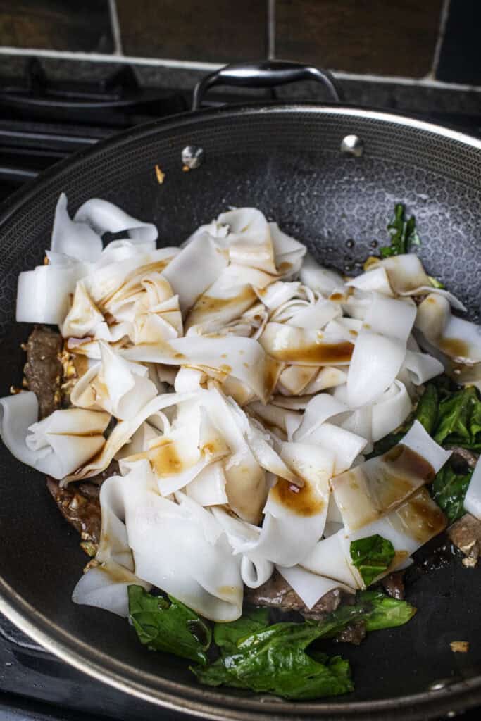 see ew noodles in a wok with greens and sauce. 