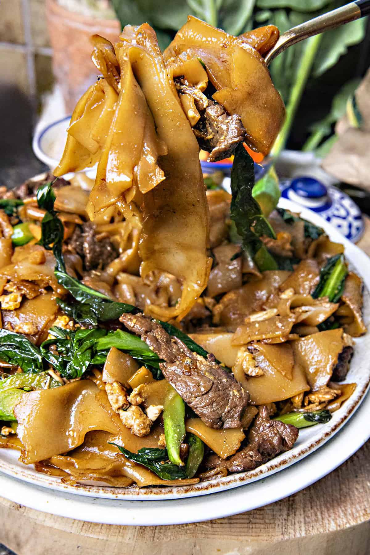 A fork, lifting a Thai pad, sees ew noodles from a plate. 