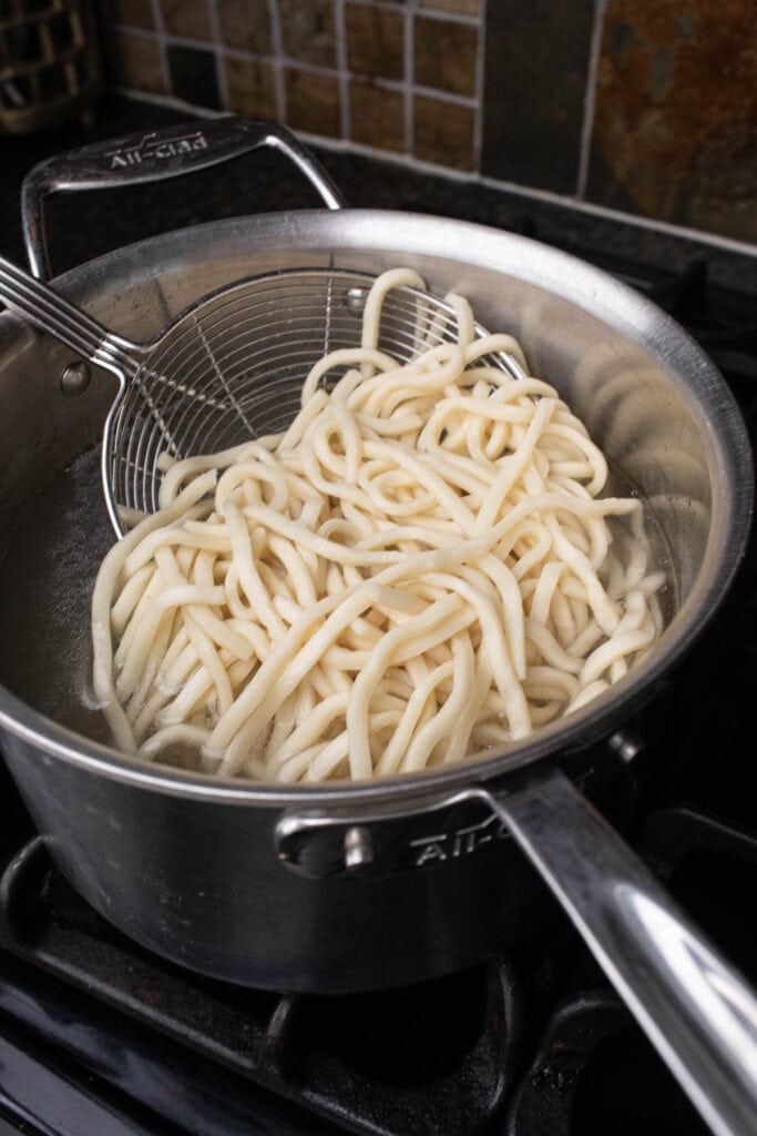 Ladle lifting udon noodles from a pot. 