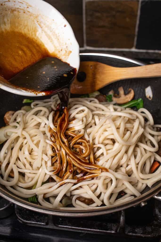 A bowl of sauce pouring over udon noodles in a wok. 