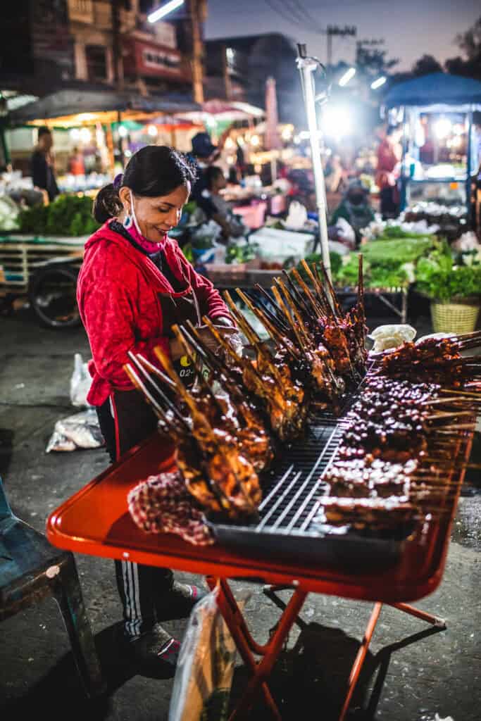 A Thai lady grilling chicken on a red table in a night market.