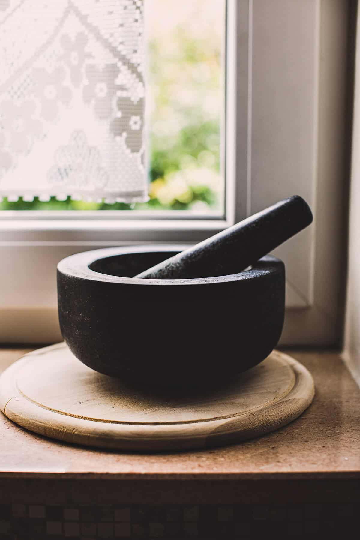 A black granite mortar and pestle on a cutting board next to a windowsill.