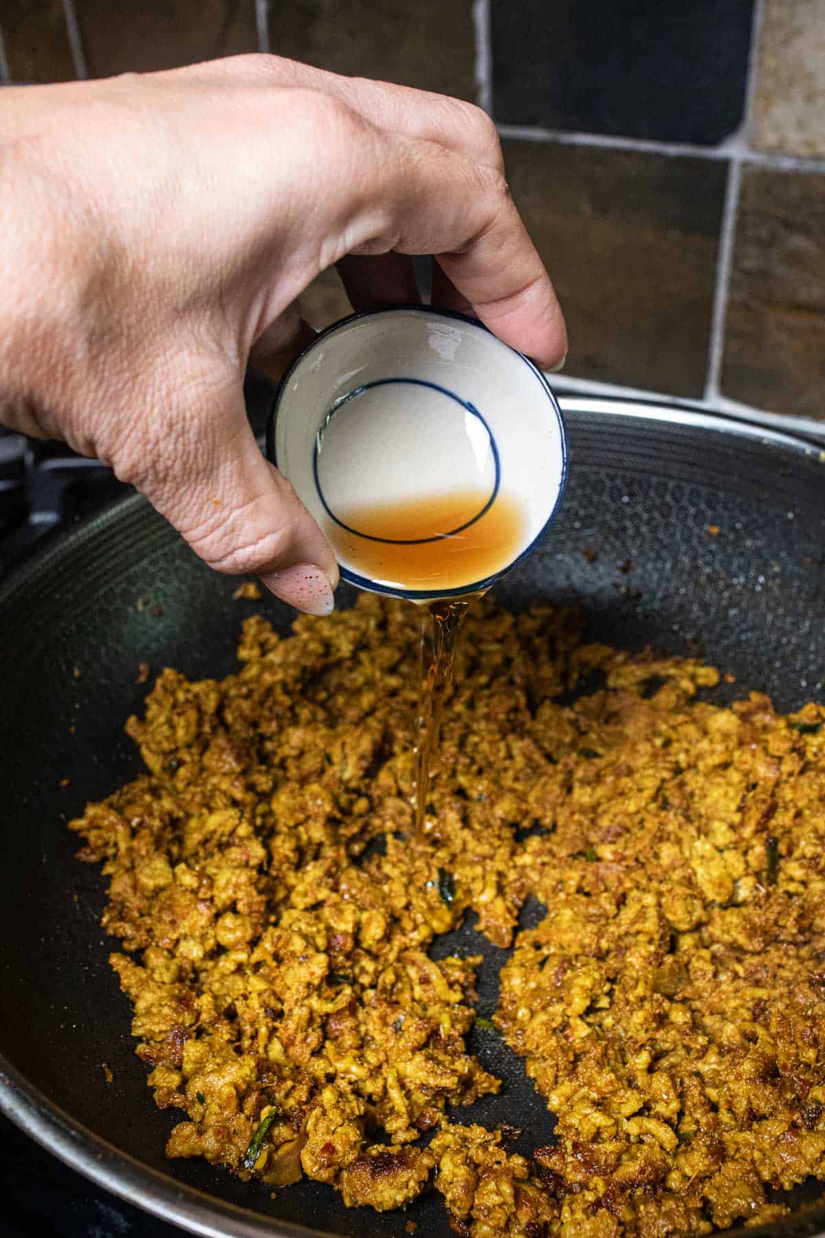 A hand holding fish sauce over minced pork curry in a wok.