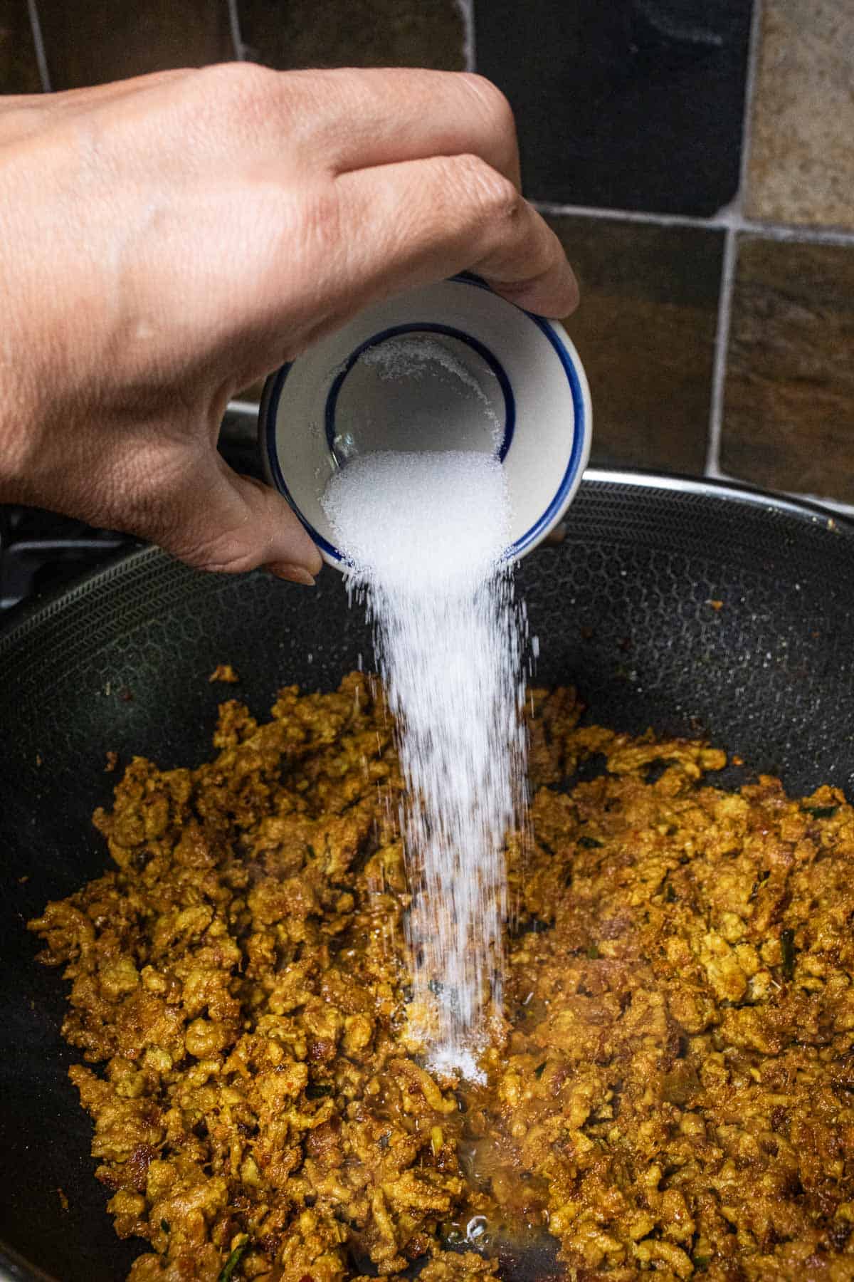 A hand holding sugar over minced pork curry in a wok.