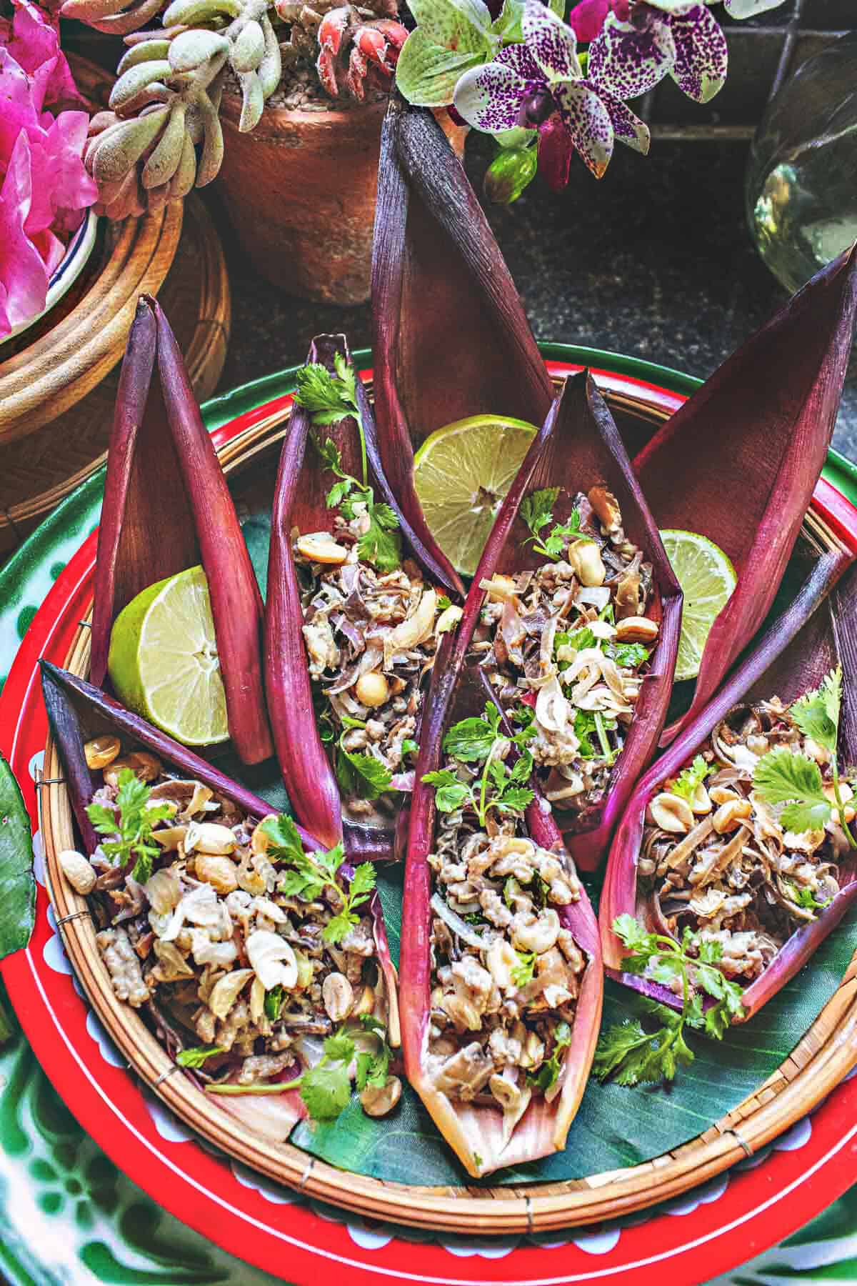 Beautiful Thai banana heart salad arranged in petal cups with lime slices and herbs, styled on a vintage green enamel tray surrounded by flowers.